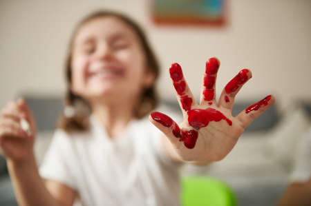 Close-up of a child's hand in red paint against a blurry smiling little girl with pigtails. Art creativityの写真素材