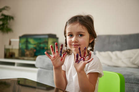Adorable little girl wearing white t-shirt looks at camera and shows her hands in paints. Art, creativityの写真素材