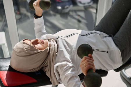 High angle view of determined Arab Muslim woman with head covered doing dumbbell bench press at home...