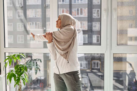 Young Arab Muslim woman stretches her arm right standing against large windows on a beautiful sunny day at home gym. Concept of home workout.の写真素材