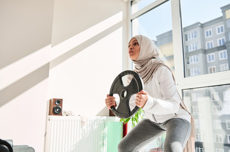 Arab muslim woman with headdress wearing hijab doing weighted squats with a metal disc in her hands...