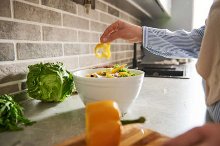 Closeup of a woman hand throwing sliced yellow pepper ring into salad bowl. Preparing vegan salad at...