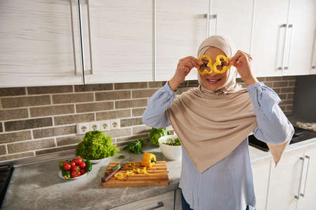 Muslim Arab woman in hijab looks through slices of yellow pepper, enjoying preparation vegan salad in the kitchenの写真素材