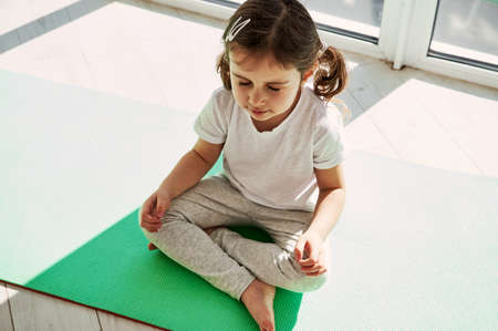 High angle view of an adorable girl with ponytails is sitting on an exercise mat and doing meditation. Developing healthy habits from early childhood.の写真素材