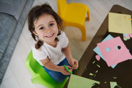 Adorable child holding scissors, cutting color paper and cute smiles to the camera. Creative hobby. Cute little girl with color paperの写真素材