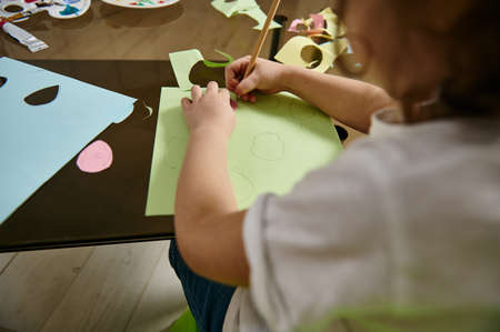 Rear view of a preschooler sitting at the table and drawing shapes on colored paper at home. Children creativity conceptの写真素材