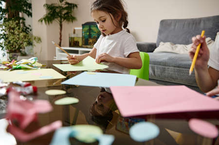 Side view to a little preschool girl sitting near her brother and drawing on a color paper