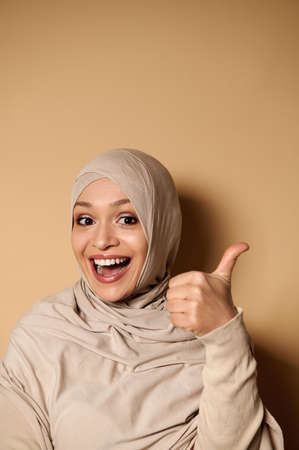 Funny woman in hijab and strict formal outfit showing thumb up and looking at camera. Beige background with copy spaceの写真素材