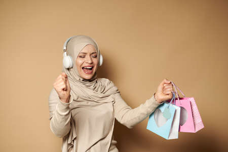 Muslim woman in hijab with headphones holding colored paper bags in her hands enjoying the upcoming religious holidays. Eid mubarak Said.の写真素材