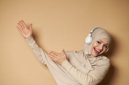 Gorgeous Muslim woman in hijab listening to the music in headphones, pointing hands on beige background with copy spaceの写真素材