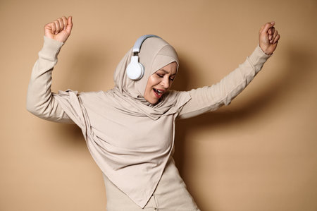 Happy Muslim woman in hijab and headphones cute smiles and relaxes, dances while listening to the music against the beige background with copy spaceの写真素材