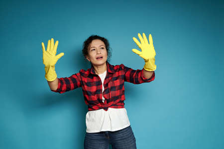Young attentive and concentrated hispanic woman looking at her hands in yellow rubber gloves for housework, pretending to touch and examine dirt on an invisible surface. Blue background with copy spaceの写真素材