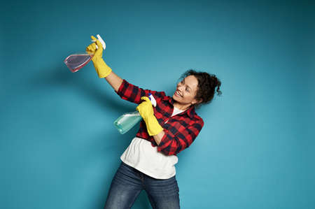 Young woman playing with cleaning sprays as if shooting a pistol. Blue background, copy space. Cleaning and home work conceptの写真素材