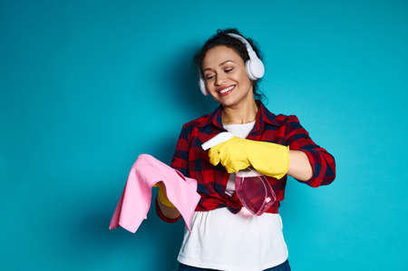Young woman with a beautiful toothy smile applying a detergent spray on a rag, cleaning concept shot on blue backgroundの写真素材
