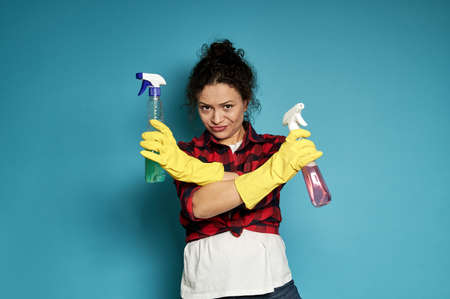Young tired exhausted disgruntled housewife crossed her arms in denial, with cleaning sprays in hands. Shot with soft shadow on blue background with copy spaceの写真素材