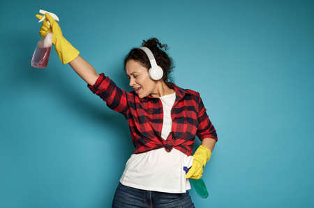 Hispanic woman, housewife, with headphones holding cleaning sprays in her hands and dancing with her eyes closed, enjoying her homework. Cleaning concept on blue background with copy spaceの写真素材