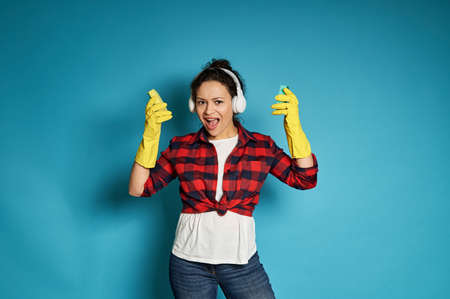 Young african woman in red plaid shirt with headphones on her head sings, posing on a blue background with cleaning sponges in her hands. Copy space.の写真素材