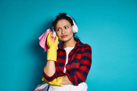 Attractive woman, housewife, standing against blue background with cleaning tools and supplies, looking intently, with a pensive look. Home work concept. Copy space.の写真素材