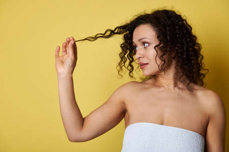 Young woman, wrapped in a bath towel, holding a lock of her curly hair and looking at her, posing against a yellow background with copy spaceの写真素材