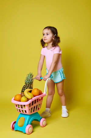 Cute little girl in pink bodysuit and blue summer shorts rolls a shopping cart full of fruits on a yellow background with copy space for advertising. Concepts, summerの写真素材