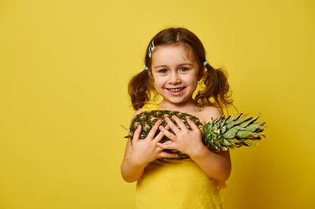 Adorable little girl with two ponytails, dressed in yellow, holding a pineapple and smiling cute, posing in front of the camera on a yellow background with copy spaceの写真素材