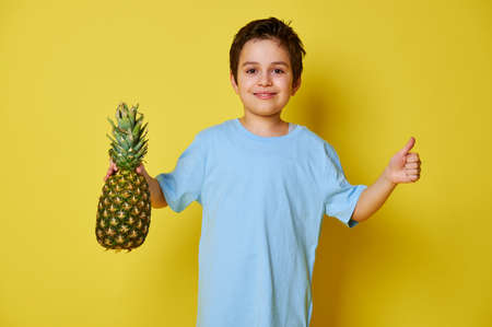 Handsome child boy holding a pineapple and showing thumb up while posing over yellow background with copy space. Summer and healthy vegan food conceptの写真素材