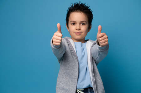 Beautiful schoolboy looking at camera and showing thumbs up while posing over blue background with space for textの写真素材