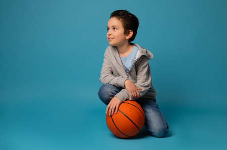 Beautiful sporty teenage boy posing with a ball for playing basketball over blue background. Concept of healthy and active lifestyle on blue background with copy spaceの写真素材