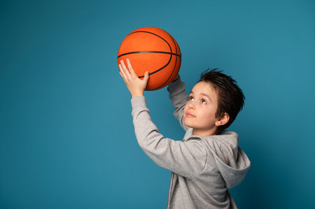 Teenage boy, basketball player, posing over blue background with an orange ball in the hands. Space...