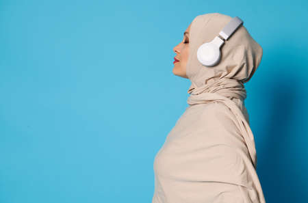 Side view of Arab Muslim woman in hijab and headphones posing over blue background with copy spaceの写真素材
