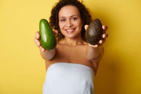 Soft focus on ripe ready-to-eat avocado fruit in hands of blurry half-naked curly woman wrapped in towel against yellow backgroundの写真素材