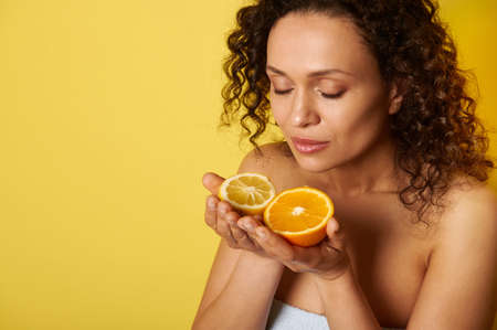 Beauty portrait of attractive curly half-naked woman with natural makeup and glowing hydrated facial skin, enjoying citrus scent in her hands. Isolated over yellow background with copy spaceの写真素材