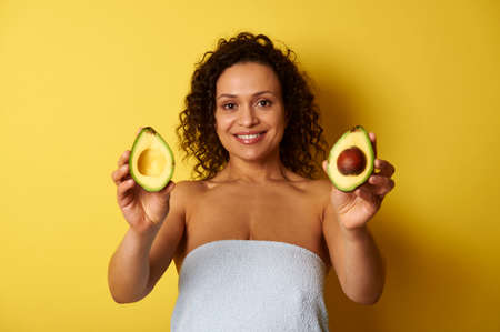 A curly-haired swarthy woman, wrapped in a bath towel holding avocado halves and showing them to camera, cute smiling with toothy smile while posing over yellow background with copy spaceの写真素材