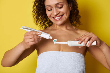 Closeup of a smiling African woman squeezing toothpaste on electric toothbrush, isolated over yellow background with copy spaceの写真素材