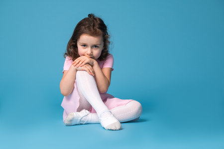 Adorable child ballerina in pink dress looking at the camera, sitting on a blue background and partially covering her mouth with her handsの写真素材