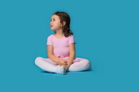 Isolated portrait of adorable girl, ballerina, sitting and looking to the side on blue background with space for textの写真素材