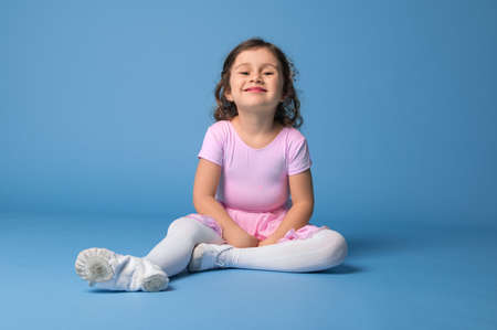 Beautiful and funny little girl, ballerina in pink dress posing to camera while relaxing on blue background with space for textの写真素材