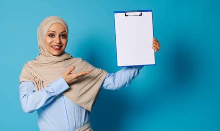A Muslim woman with a covered head in a hijab holds a clipboard in one hand and points at it with her hands, presenting invisible information on a blank sheet of paper. Isolated over blue backgroundの写真素材