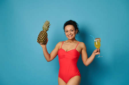 Attractive fit woman in red swimsuit posing against blue background with a glass of cocktail and a pineapple in her handsの写真素材