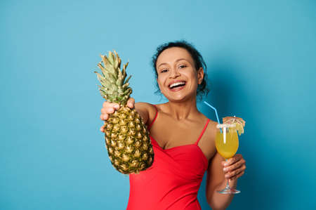 Charming brunette with beautiful smile holding a glass with cocktail and showing a pineapple to the camera. Summer concepts with space for textの写真素材