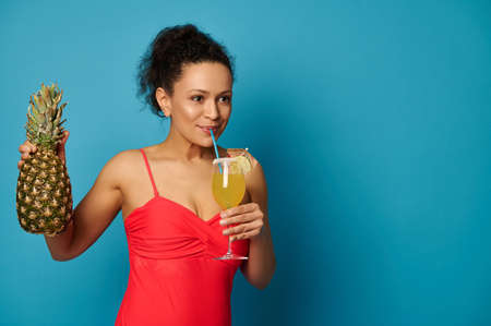 Young woman with a pineapple drinking a delicious exotic cocktail from a straw, posing over blue...