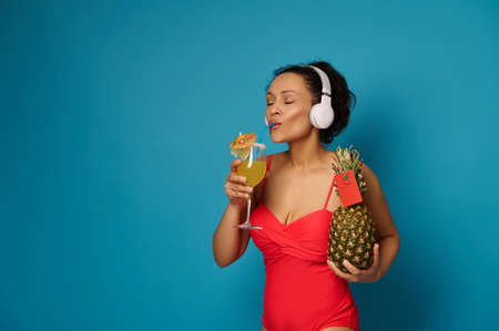 Mixed race woman in red swimsuit holds pineapple with blank red card and drinks exotic cocktail while enjoying music with headphones. Summer concepts on blue background with copy spaceの写真素材