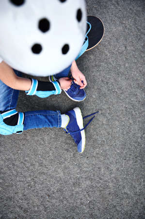 High angle view of unrecognizable boy in safety helmet for skateboard tying shoelaces sitting on asphalt of playgroundの写真素材