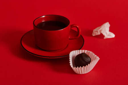 Closeup of a ceramic coffee cup with coffee in a red saucer, next to chocolate truffle and a crumpled paper wrapper from a chocolate praline. Coffee and chocolate day conceptsの写真素材