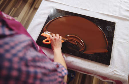 Top view of chocolatier holding cake scraber and cooling melted chocolate mass on a marble table....