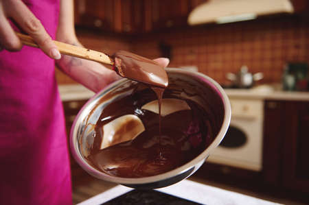 Pastry chef manufacturer of chocolate confectionery mixing melted chocolate mass in a steel bowl with a wooden spoon. Manufacturing homemade chocolate candies for celebration of worlds chocolate dayの写真素材