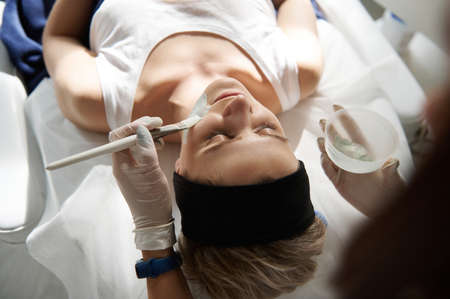 Overhead view of young woman relaxing on a massage table during beauty spa procedure and doctor cosmetologist applying alginate mask on her face. Professional skin care concept in SPA salonの写真素材