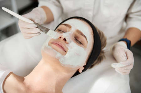 Close-up portrait of a beautician's hand applying a beauty mask on the face of a young woman with perfect clean skin with a brush. Professional skin care concept in modern beauty spa salonsの写真素材