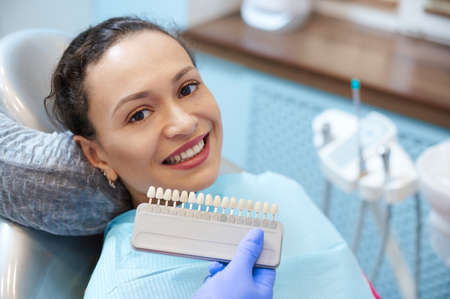 Brunette woman with beautiful smile before receiving dental care check up and teeth whitening bleaching, female doctor wearing exam gloves checks tooth color with a comparison veneer scale chart.の写真素材