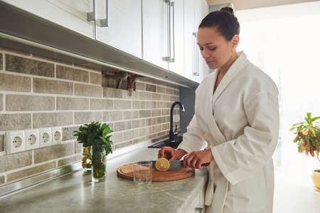 Charming woman in white bathrobe standing in the home kitchen and cutting lemon for preparing lemon water , healthy drink in the early morningの写真素材